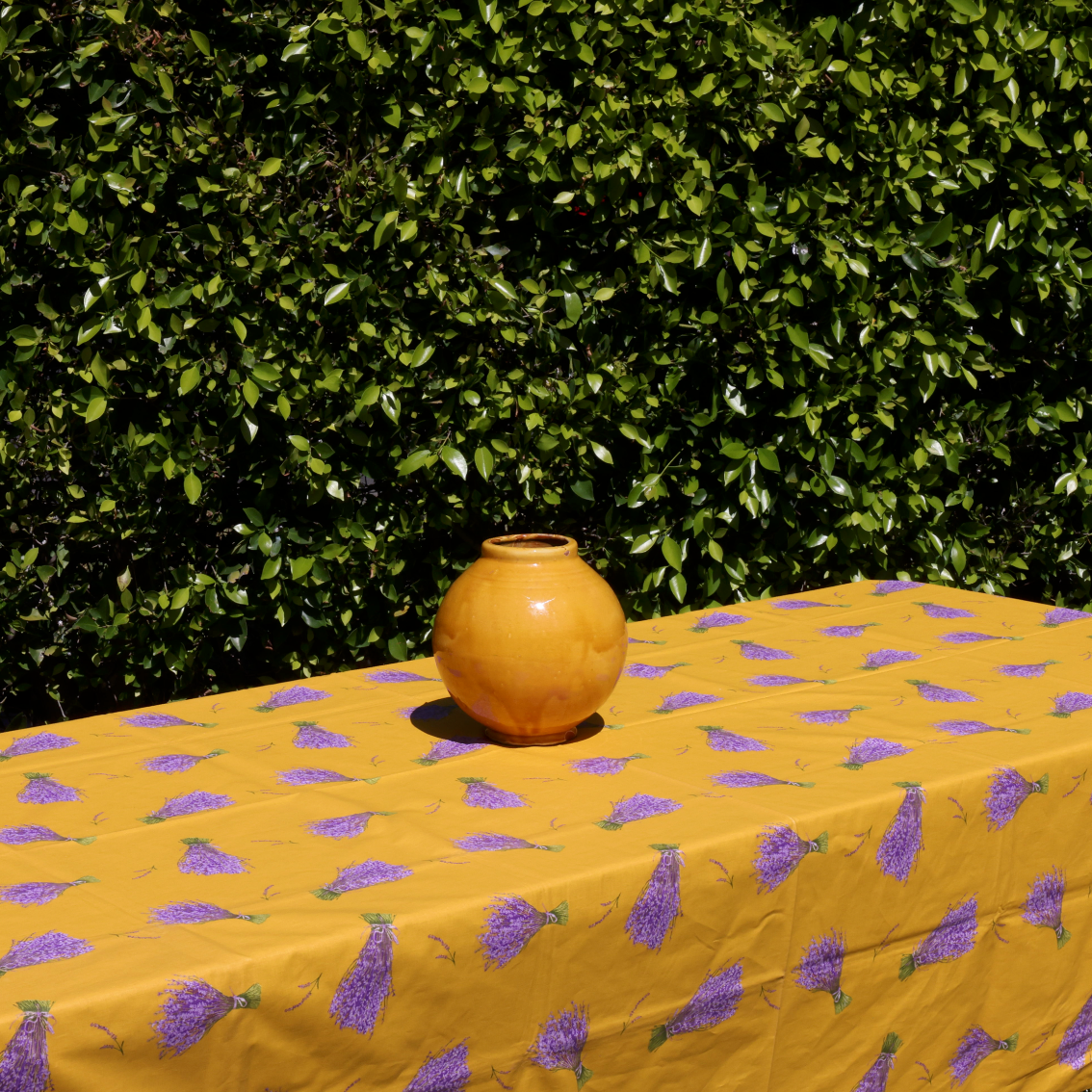 Yellow tablecloth with purple floral pattern and a brown vase on a green hedge background
