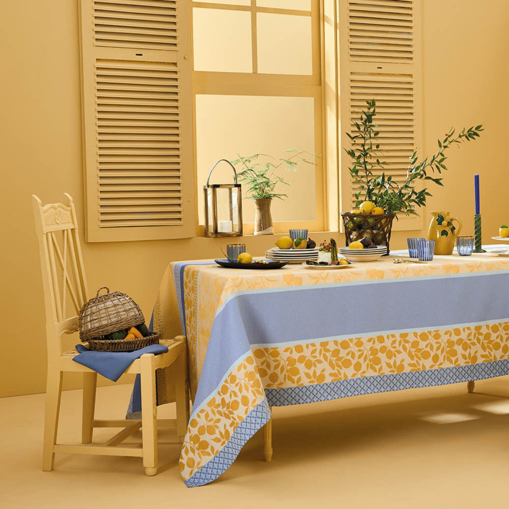 Mille Citrons Provence Jacquard Tablecloth in blue and yellow cotton with lemon motifs, styled on a dining table with matching place settings in a warm yellow room.