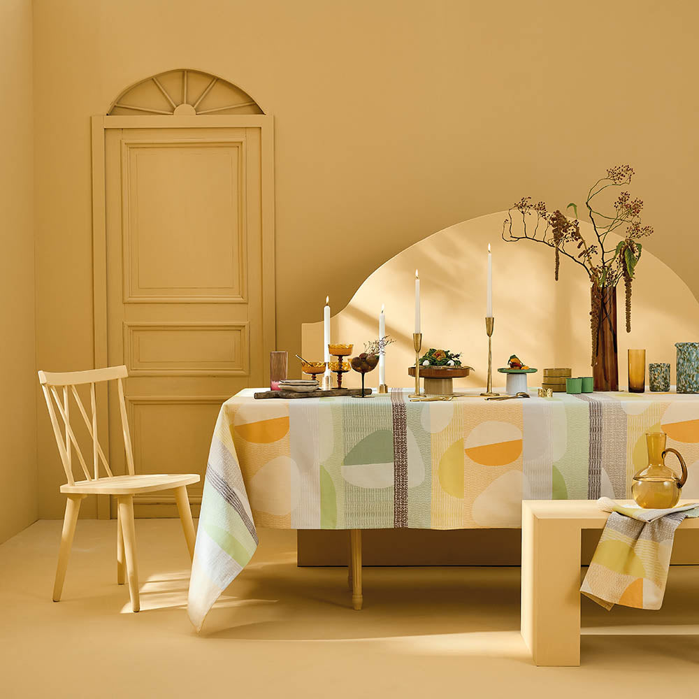 Mille Galets Terre Cuite Tablecloth, cotton jacquard with yellow, brown, and green circular pebble motif, set for a formal dining scene in a warm yellow room.