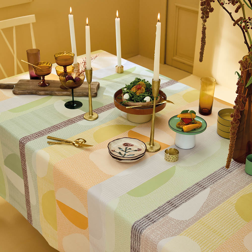 Mille Galets Terre Cuite Tablecloth, cotton jacquard with yellow, brown, and green circular pebble motif, on a table set with candles, and mix matched dinnerware.