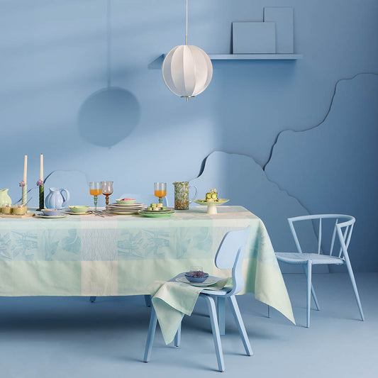 Mille Iris D'Eau Aqua Tablecloth, blue and white iris floral madras plaid on cotton jacquard, set for a garden-inspired dining scene in a sky blue room and matching chairs.