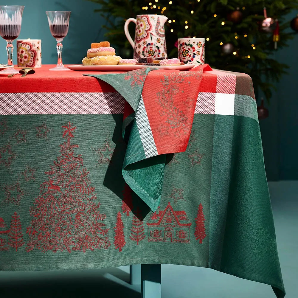 A holiday table setting featuring the Garnier-Thiebaut Noel Magique Houx green and red cotton jacquard tablecloth and matching napkin in a warm teal room with Christmas decorations.