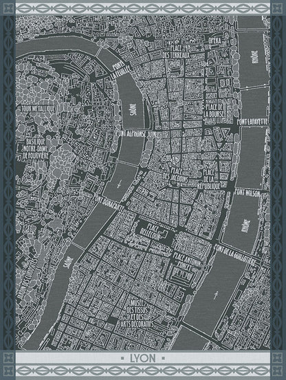 A dark gray and white street map print of Lyon, France, on a cotton dishtowel featuring labeled landmarks, rivers, and roads, framed by a decorative border with the word 'LYON' at the bottom.