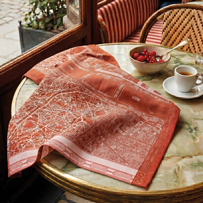 A red dishtowel with a white map print of Paris, draped over a round green marble table in a French cafe setting next to a bowl of cherries and a cup of coffee.