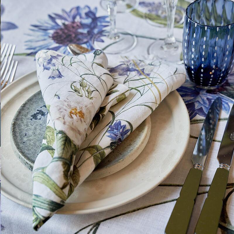 Table setting with a folded Blue Flower Garden linen napkin on a plate, and cutlery on a matching tablecloth.