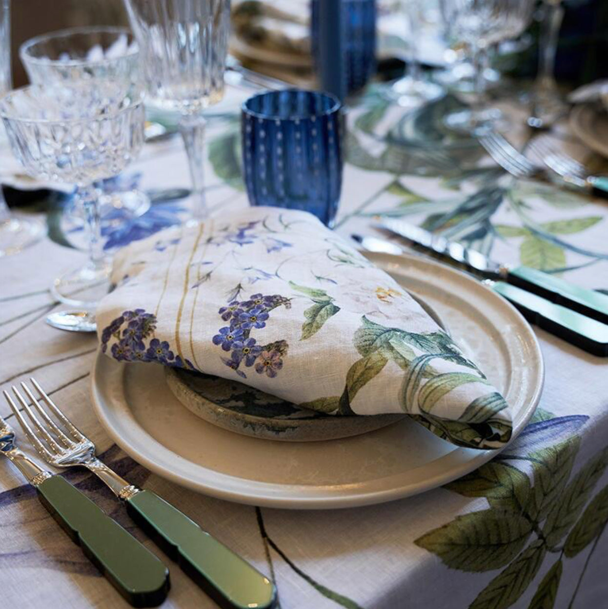 Dining table setting with a folded Blue Garden linen napkin, plates, and cutlery on a matching tablecloth.