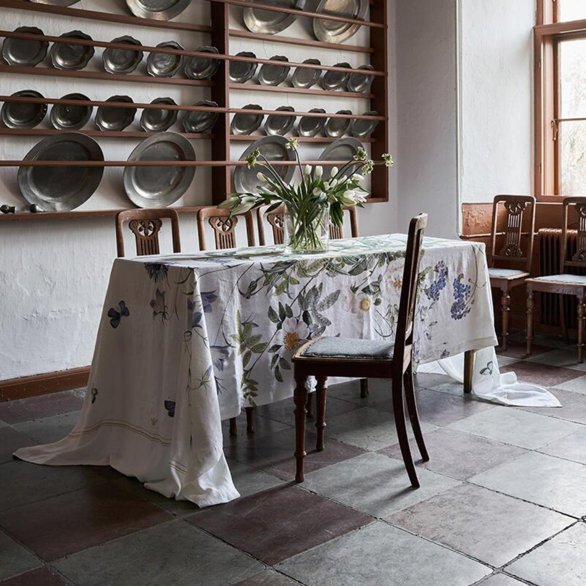 An elegant dining room featuring a long, flowing floral linen tablecloth with blue bellflowers, white roses, and butterflies, draped over a rustic table with classic wooden chairs.