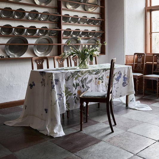 An elegant dining room featuring a long, flowing floral linen tablecloth with blue bellflowers, white roses, and butterflies, draped over a rustic table with classic wooden chairs.