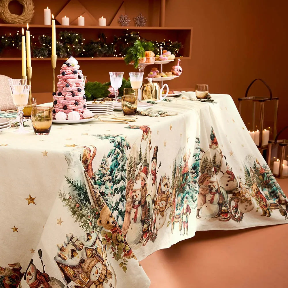 A festive holiday dining table dressed with a cream cotton-linen tablecloth featuring a vintage print of snowmen, Santas, and evergreen trees, styled alongside an elegant pink macaron tower and glowing candlesticks against a warm terracotta background.