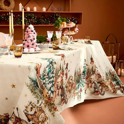 A festive holiday dining table dressed with a cream cotton-linen tablecloth featuring a vintage print of snowmen, Santas, and evergreen trees, styled alongside an elegant pink macaron tower and glowing candlesticks against a warm terracotta background.