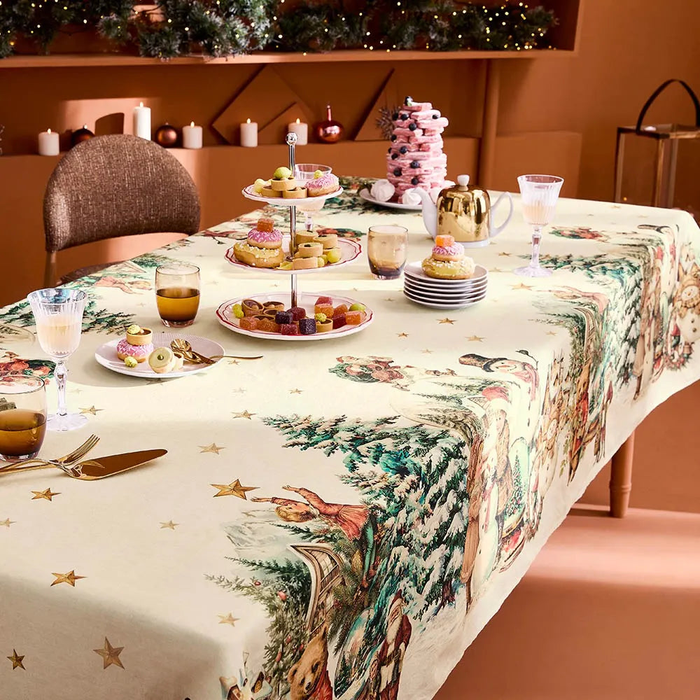 A festive holiday dining table dressed with a cream cotton-linen tablecloth featuring a vintage print of snowmen, Santas, and evergreen trees, styled alongside an elegant pink macaron tower and glowing candlesticks against a warm terracotta background.