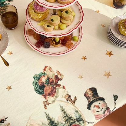 Closeup of a festive holiday cream cotton-linen tablecloth featuring a vintage print of snowmen, Santa, and gold stars with holiday plates of treats. 