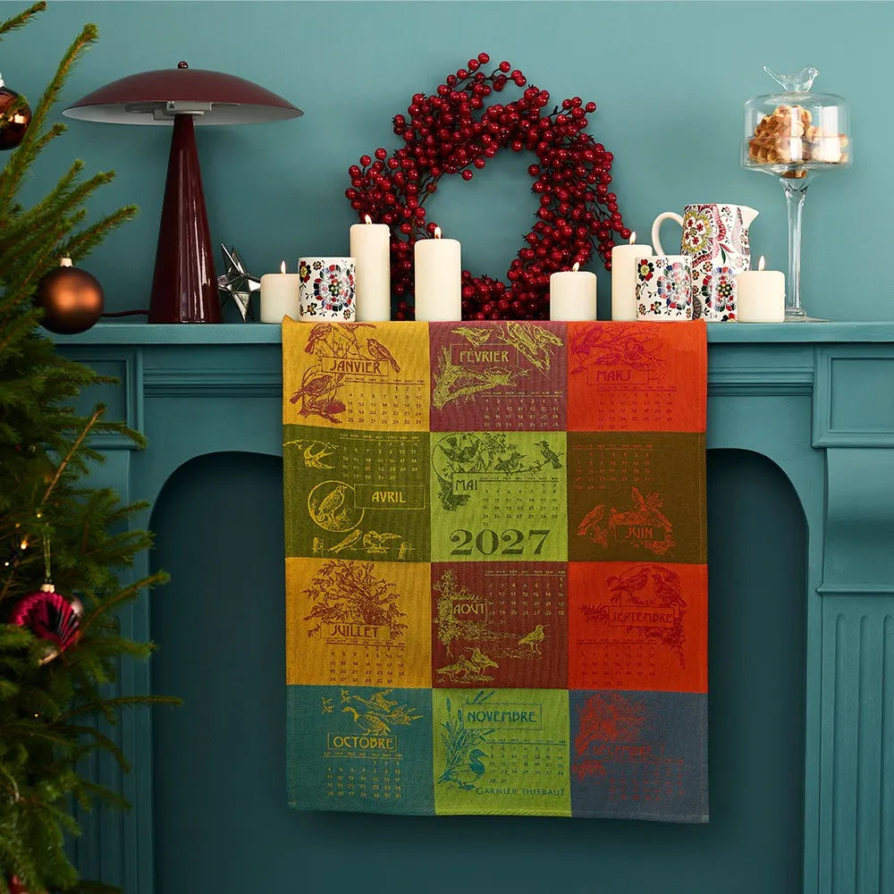 A square patchwork 2027 calendar dishtowel with printed month panels in yellow, green, orange, and red, draped over a teal fireplace mantel decorated with candles, a red berry wreath, mugs, and a Christmas tree.