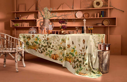 A long table draped with a cream tablecloth featuring a detailed autumn harvest pattern of pumpkins, gourds, leaves, and flowers, styled in a warm, rustic room with wooden shelves and a white spindleback chair.