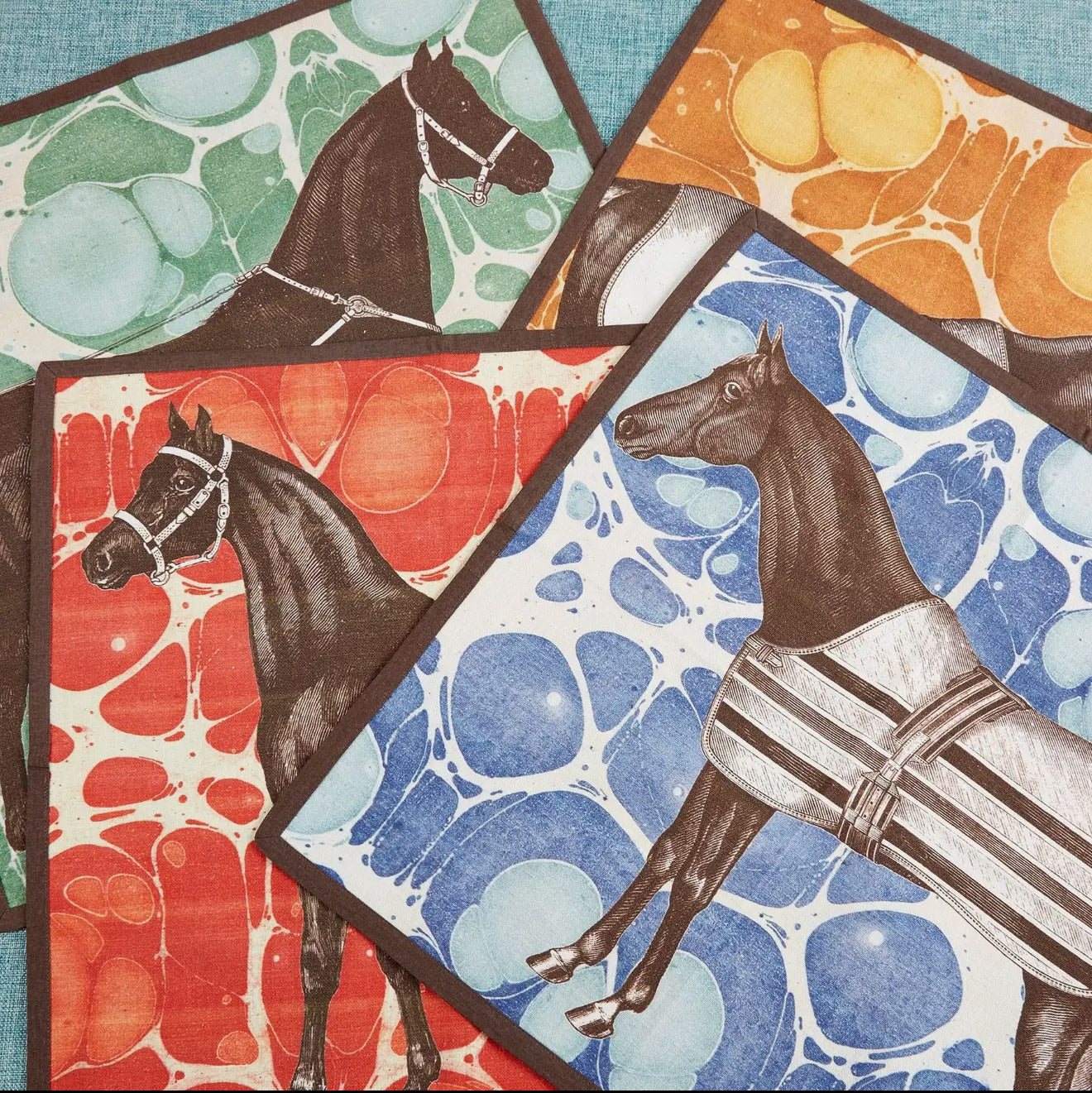 Set of four Horse Marble cotton, square napkins arranged on a blue background. Each square napkin features a unique, elegantly illustrated brown horse motif set against a vibrant, marbled background pattern in shades of yellow gold, green, red, and blue, all finished with a crisp, dark brown piped edge.