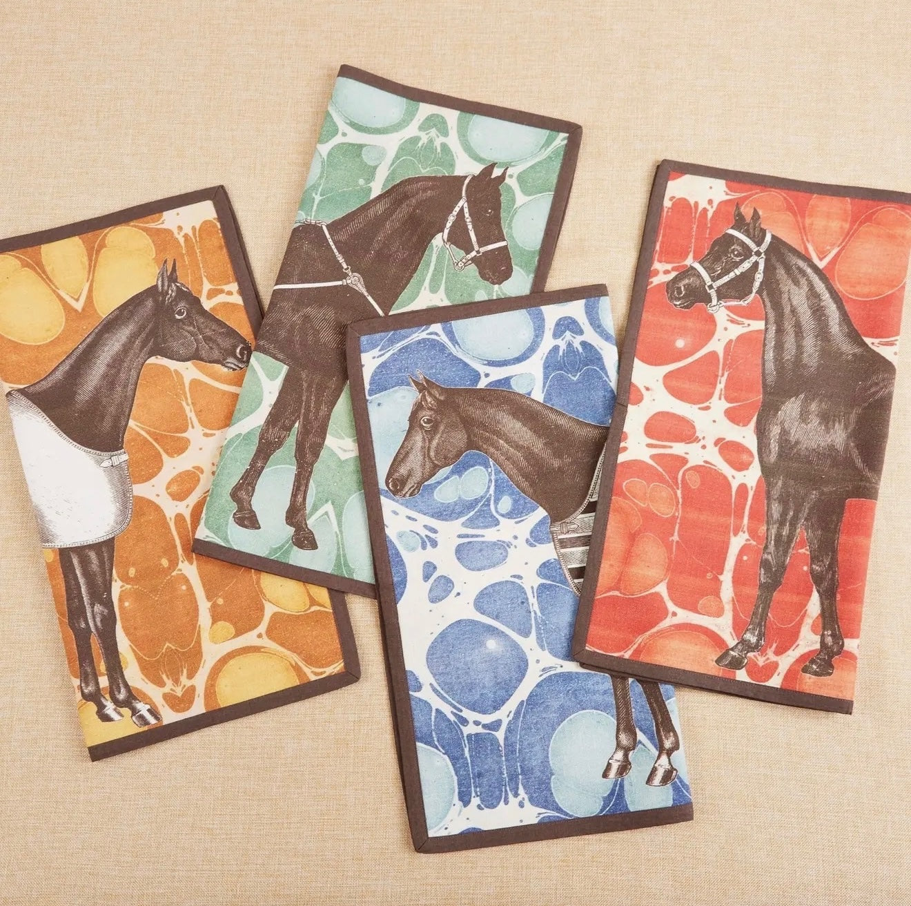 Set of four Horse Marble cotton dinner napkins arranged on a neutral fabric background. Each square napkin features a unique, elegantly illustrated brown horse motif set against a vibrant, marbled background pattern in shades of yellow gold, green, red, and blue, all finished with a crisp, dark brown piped edge.