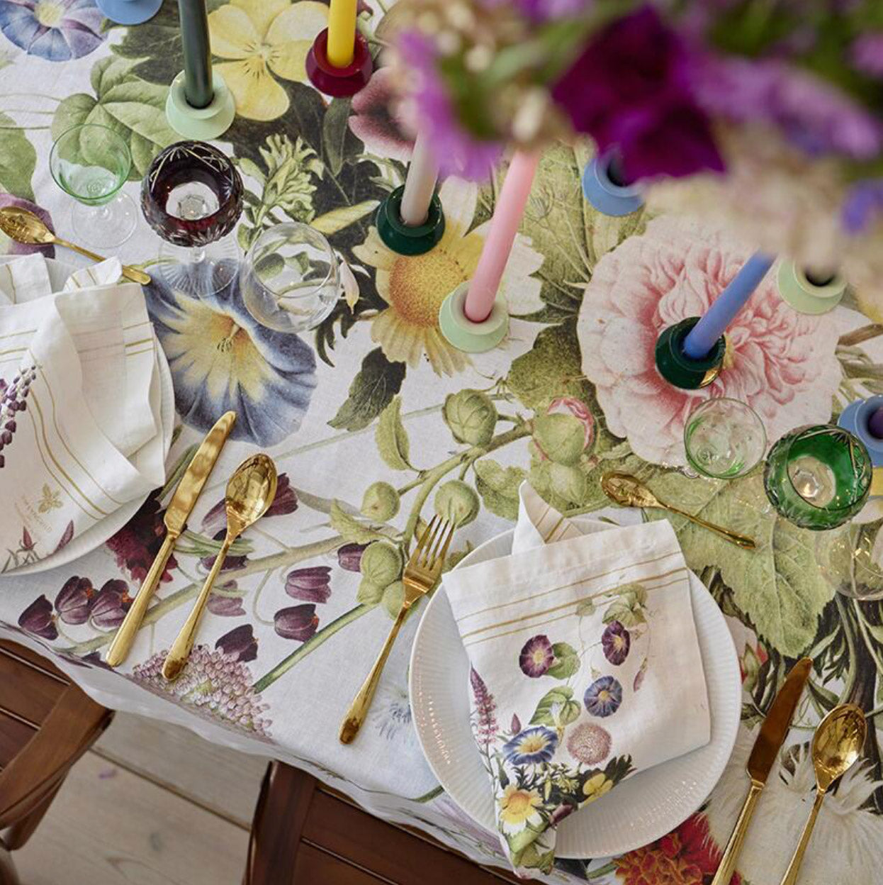 Decorative table setting with floral tablecloth, cutlery, and candles on a wooden table.