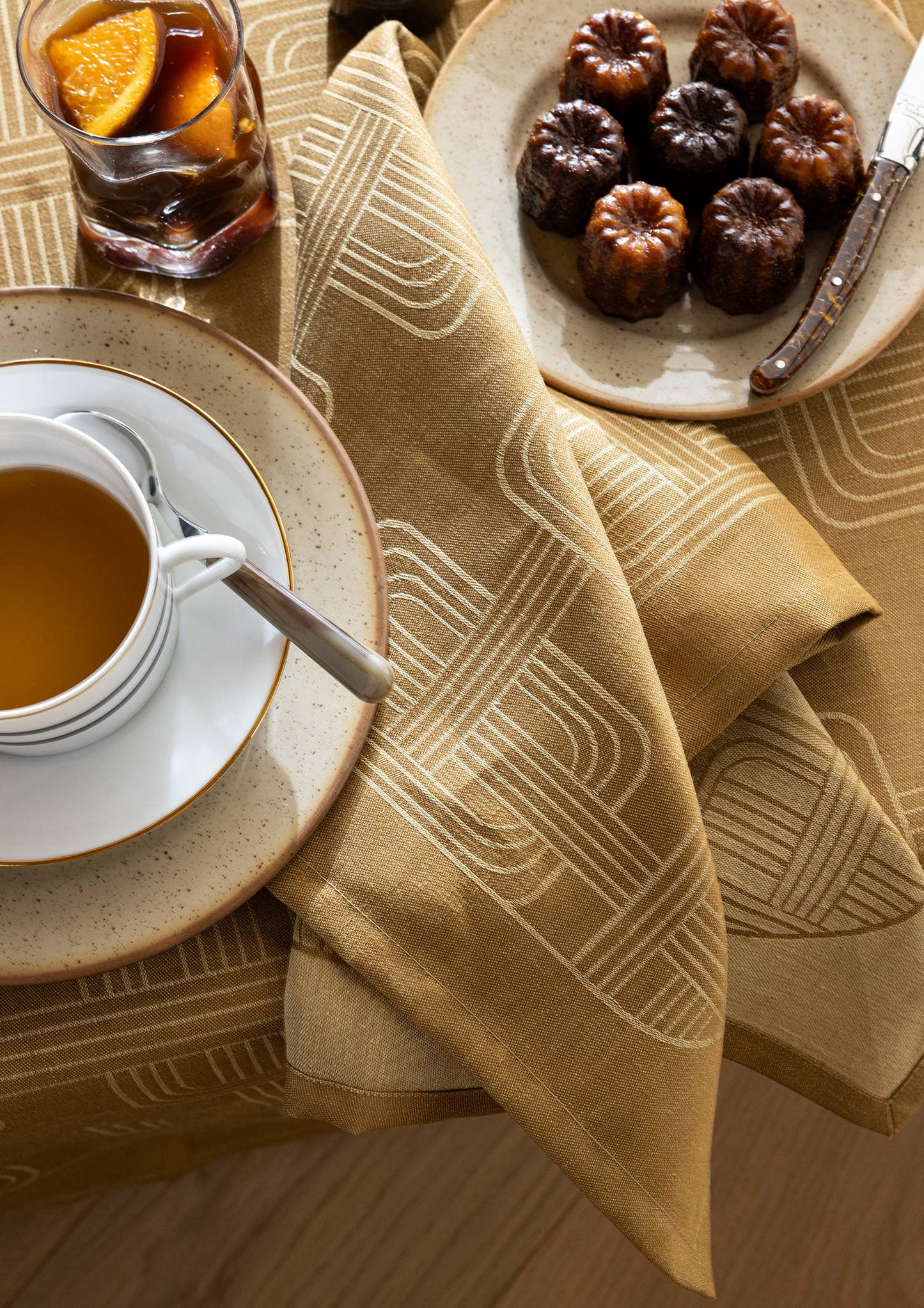 Close-up tabletop view of a brown Hôtel Particulier Jacquard linen napkin draped over a matching tablecloth, set beside a tea cup, a plate of mini cannelé pastries, and iced tea.