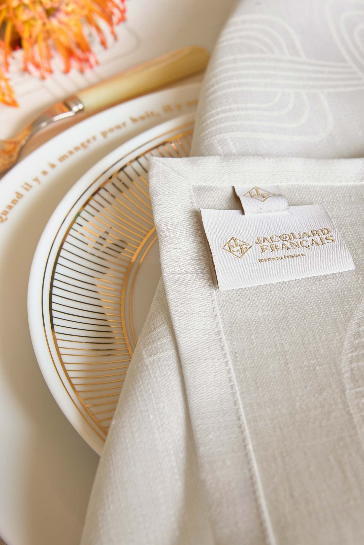 Close-up shot of a folded light grey Jacquard linen napkin with a visible Le Jacquard Français woven brand label, styled over a gold-rimmed white plate next to a wooden spoon and orange flowers.