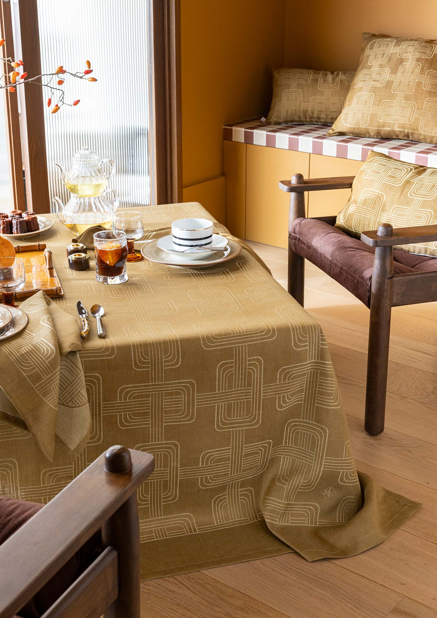 Dining table draped in a brown Hôtel Particulier Jacquard linen tablecloth with a geometric link pattern, set for tea with white cups, clear glasses, and a glass teapot in a warm room setting.
