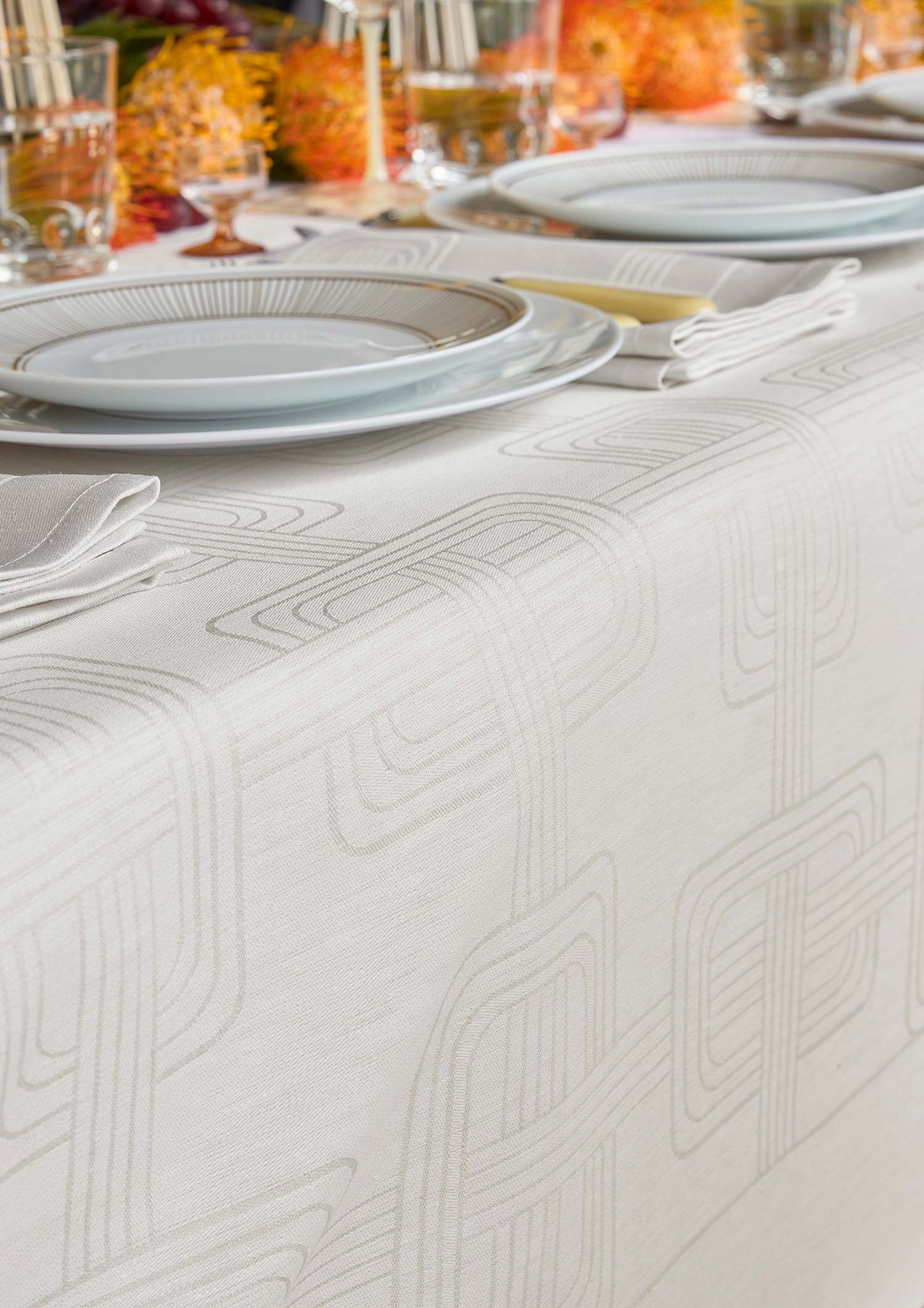 Close-up view of a light grey Hôtel Particulier Jacquard linen tablecloth styled on a dinner table with layered white plates, folded napkins, and warm orange floral decorations in the background.
