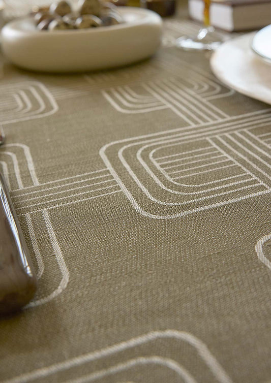 A close-up of the Hôtel Particulier woven tablecloth in a warm green color with a white geometric pattern of nested rectangles and lines. The table is styled with dining props including a plate, a glass, and a bowl in a softly blurred background.