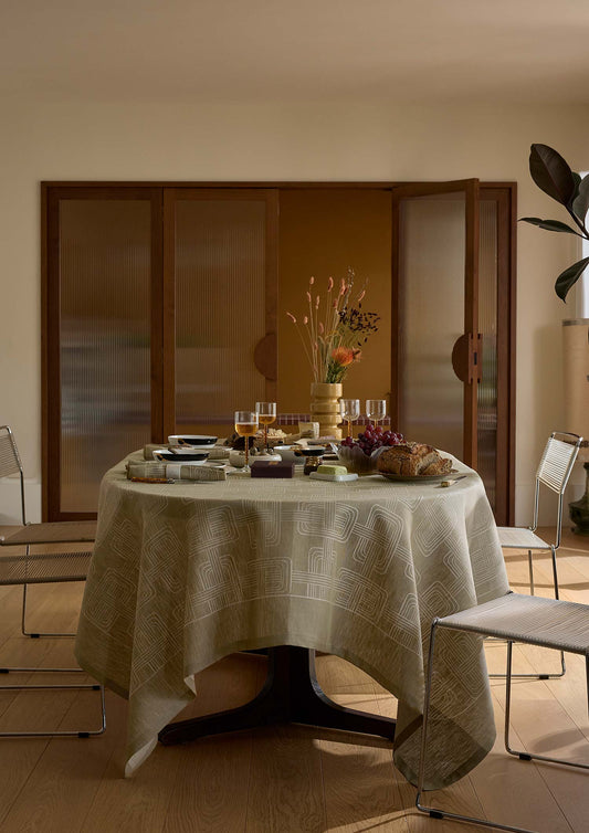 A round dining table covered with a beige patterned tablecloth, set with glassware, plates, and a floral centerpiece in a warm, modern dining room.