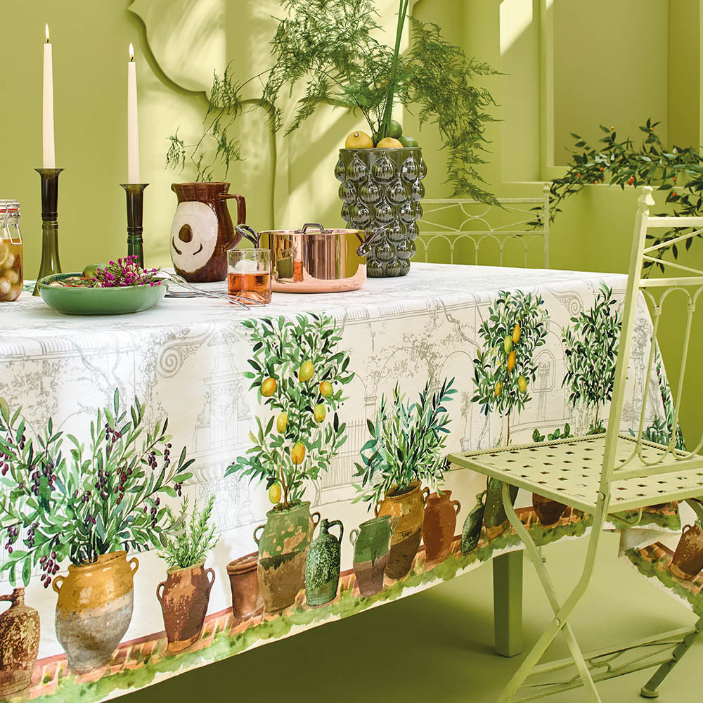 Jardin Provencal Olive Tablecloth made with cotton-linen blend covers a dining table featuring potted plants, candles, and a vase.