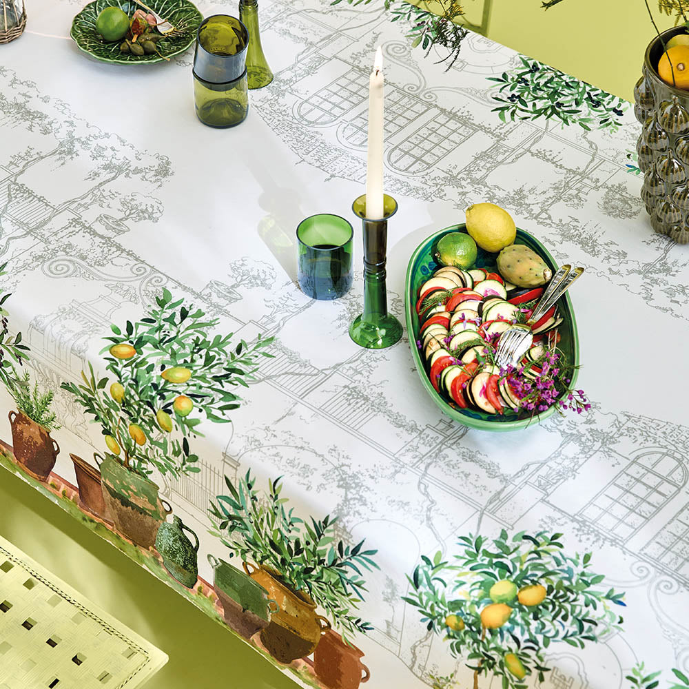 Jardin Provencal Olive Tablecloth with garden-themed design featuring a hand sketch of a French village and potted plants underneath lemons on a table setting.