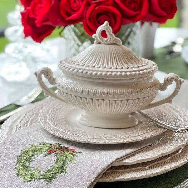 An off-white ceramic lidded tureen with ornate relief detailing, shown on a matching plate and folded linen napkin in a styled table setting with red roses in the background.