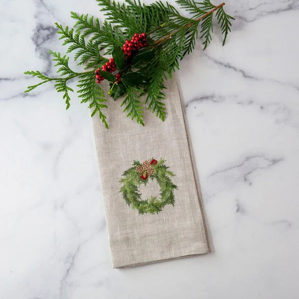 A folded flax colored linen napkin with an embroidered green wreath and red berries design, styled with a pine branch on a white marble surface.