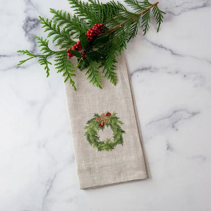 A folded flax colored linen napkin with an embroidered green wreath and red berries design, styled with a pine branch on a white marble surface.