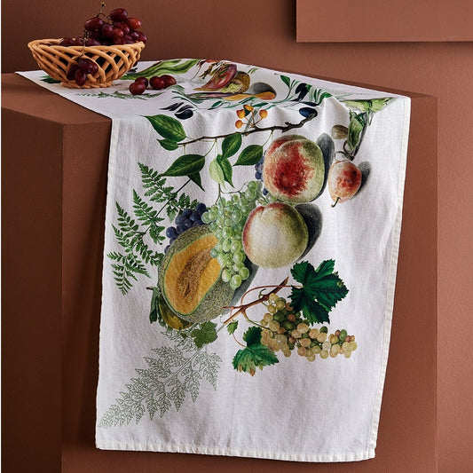 A close-up view of a cream cotton-linen tablecloth styled as a runner over a modern terracotta block table, featuring a vibrant printed harvest illustration of ripe melons, green and purple grapes, peaches, and lush green ferns.