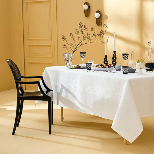 Dining table set with Le Cercle Blanc Jacquard stain-resistant tablecloth, black chairs, and decorative items in a warm-toned room.