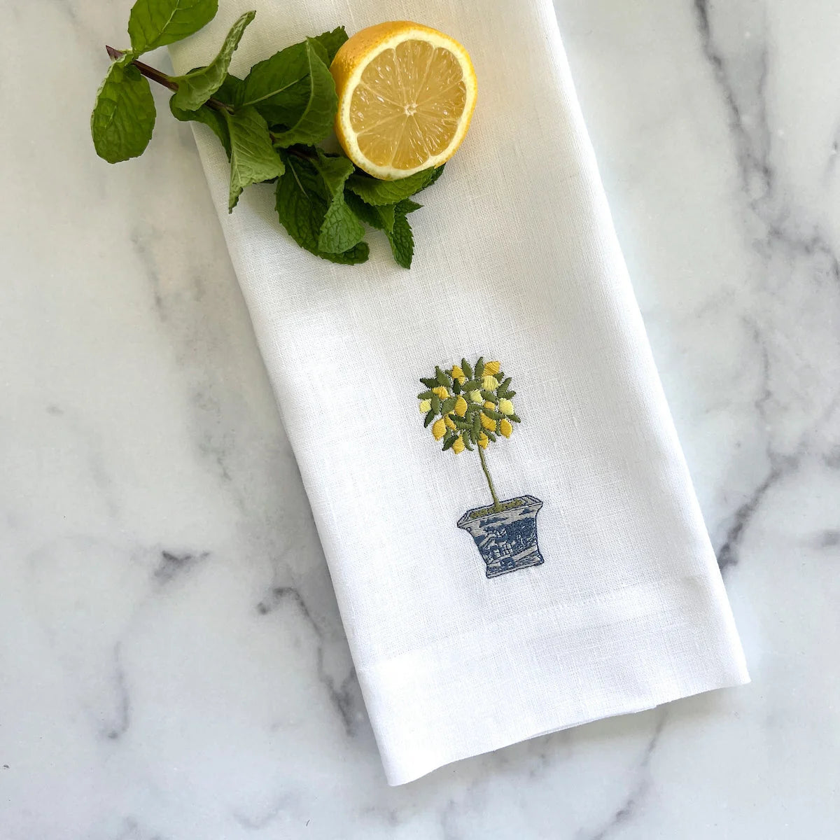 A white linen kitchen towel with an embroidered lemon tree in a pot, styled with a fresh lemon half and mint leaves on a white marble surface.