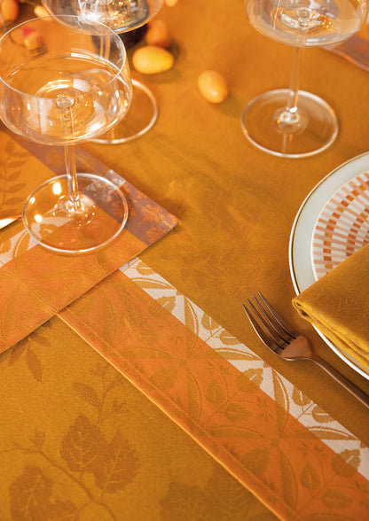 Detailed shot of the Manor Lane orange-gold Jacquard tablecloth and placemat showing the woven leaf border design, mitered edge, and table stemware.