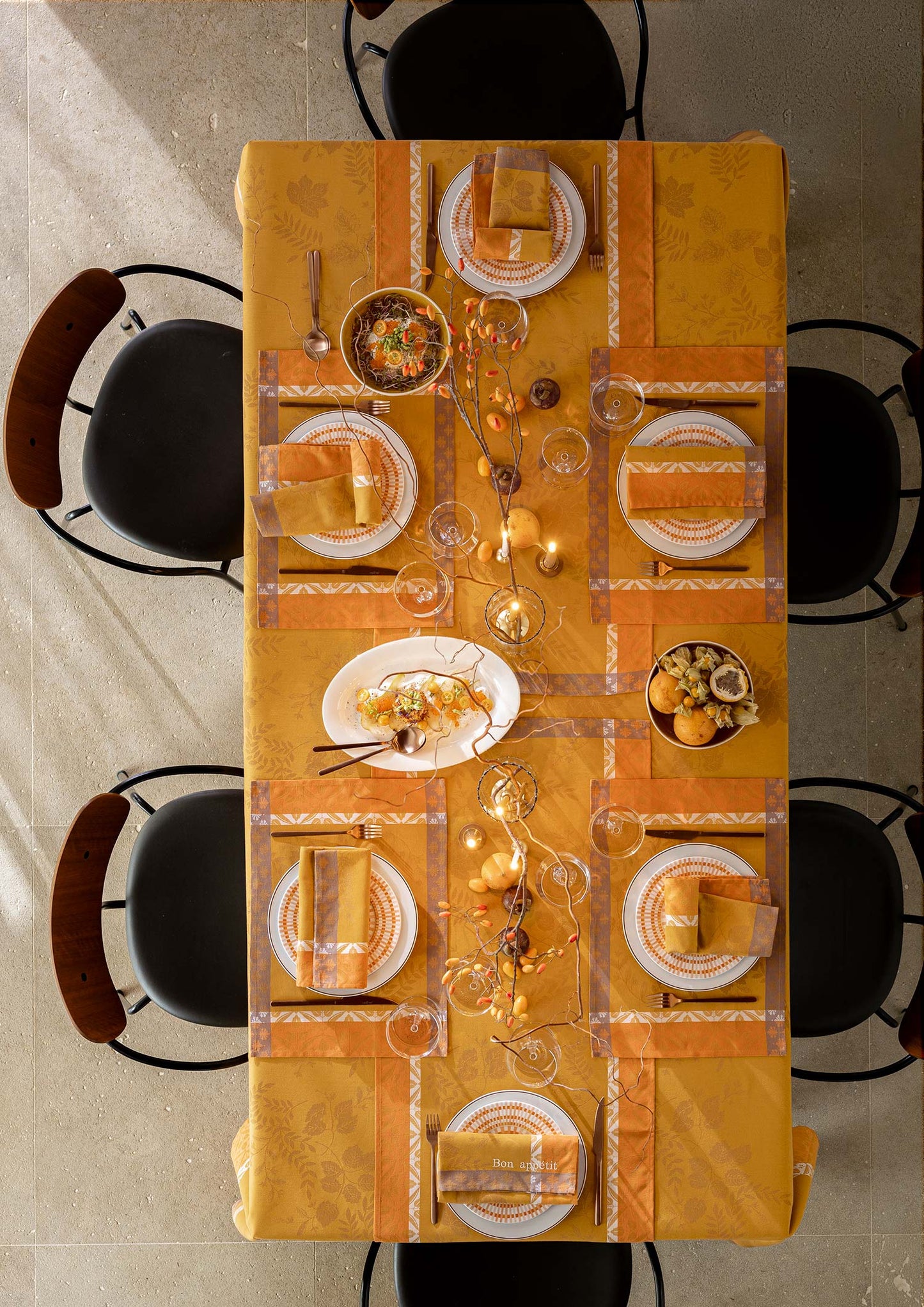 Overhead view of an autumn dining table setting featuring a mustard yellow Jacquard tablecloth, white place settings, folded napkins, and gold-toned glassware surrounded by dark chairs.