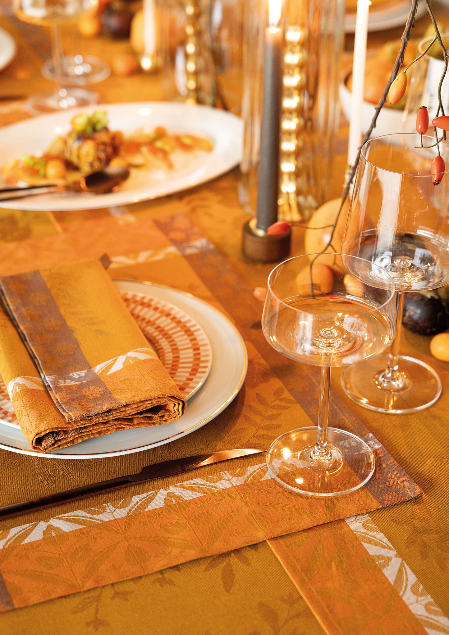 Close-up view of a fall dining table set with an orange-gold Manor Lane Jacquard tablecloth, a folded cloth napkin on a patterned plate, two clear wine glasses, and a tall candle.