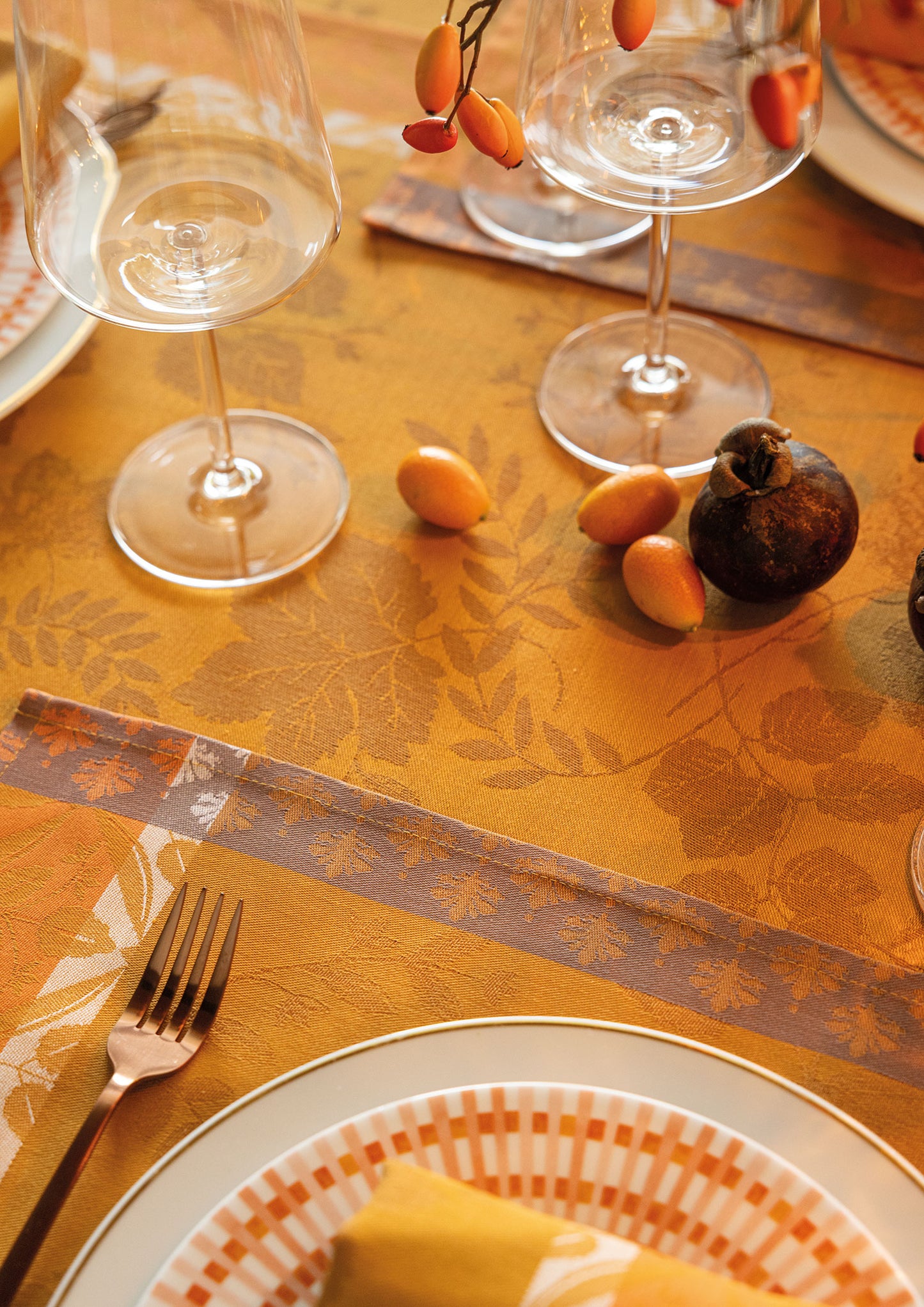 Close-up view of an orange-gold Manor Lane Jacquard tablecloth set with a white grid-patterned dinner plate, a gold fork, two clear wine glasses, and decorative rose hips and a fig on the table.
