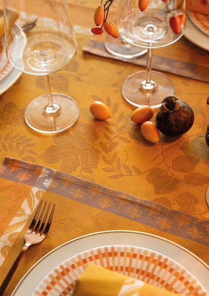 Close-up view of an orange-gold Manor Lane Jacquard tablecloth set with a white grid-patterned dinner plate, a gold fork, two clear wine glasses, and decorative rose hips and a fig on the table.
