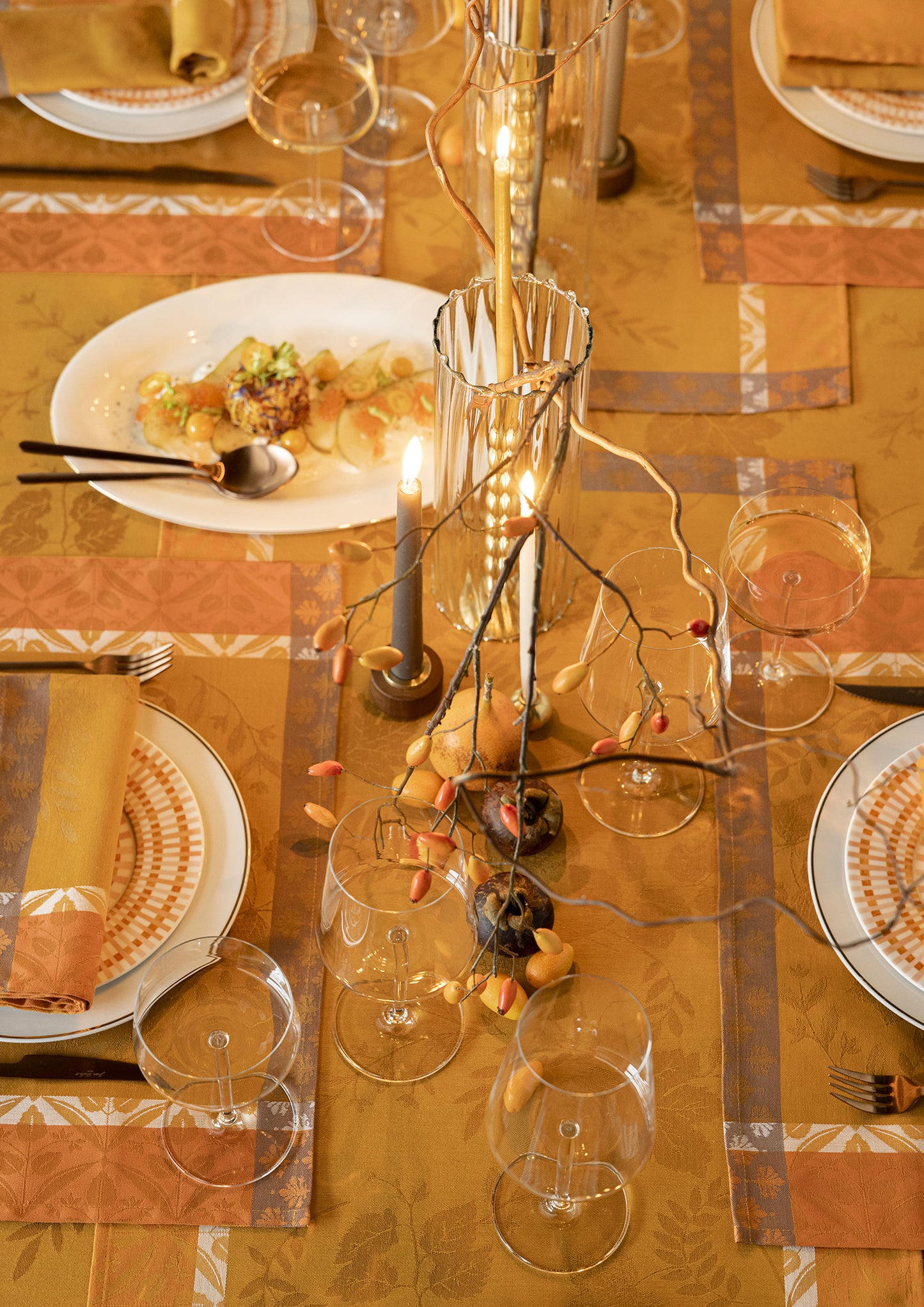 Top-down view of an autumn table setting on a mustard yellow Jacquard tablecloth, featuring wine glasses, candlelit glass holders, dark blue candles, white dinnerware, and decorative rose hips.