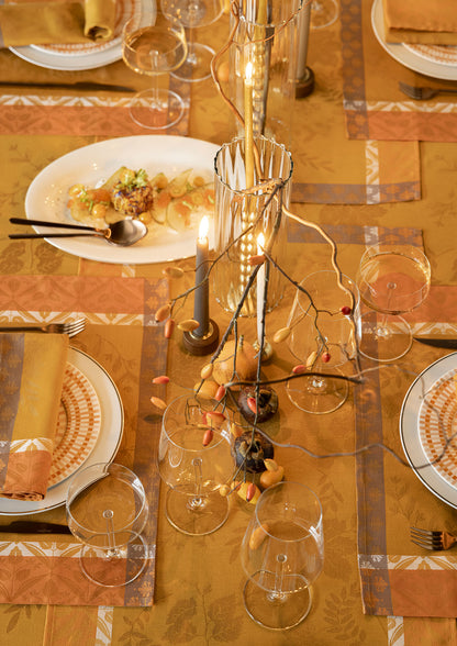 Top-down view of an autumn table setting on a mustard yellow Jacquard tablecloth, featuring wine glasses, candlelit glass holders, dark blue candles, white dinnerware, and decorative rose hips.