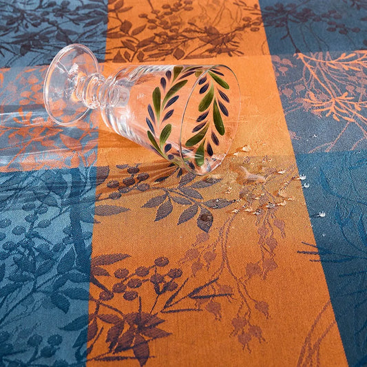 A close-up view of a luxury coated jacquard tablecloth featuring an intricate orange and deep teal autumn leaf pattern, with an elegant clear wine glass and spilled water.