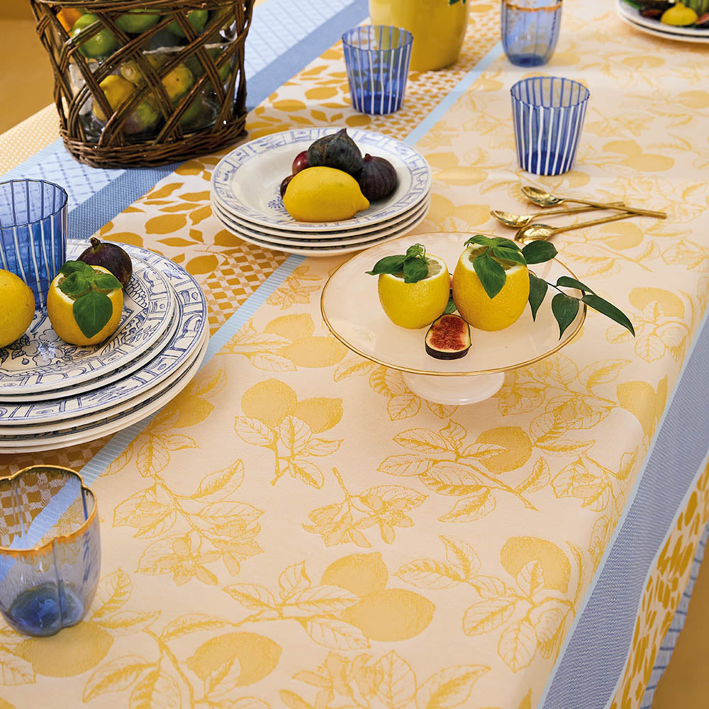 Mille Citrons Provence Jacquard Tablecloth in stripes of blue and yellow cotton with lemon motifs, styled on a dining table with a matching place settings and fruits.