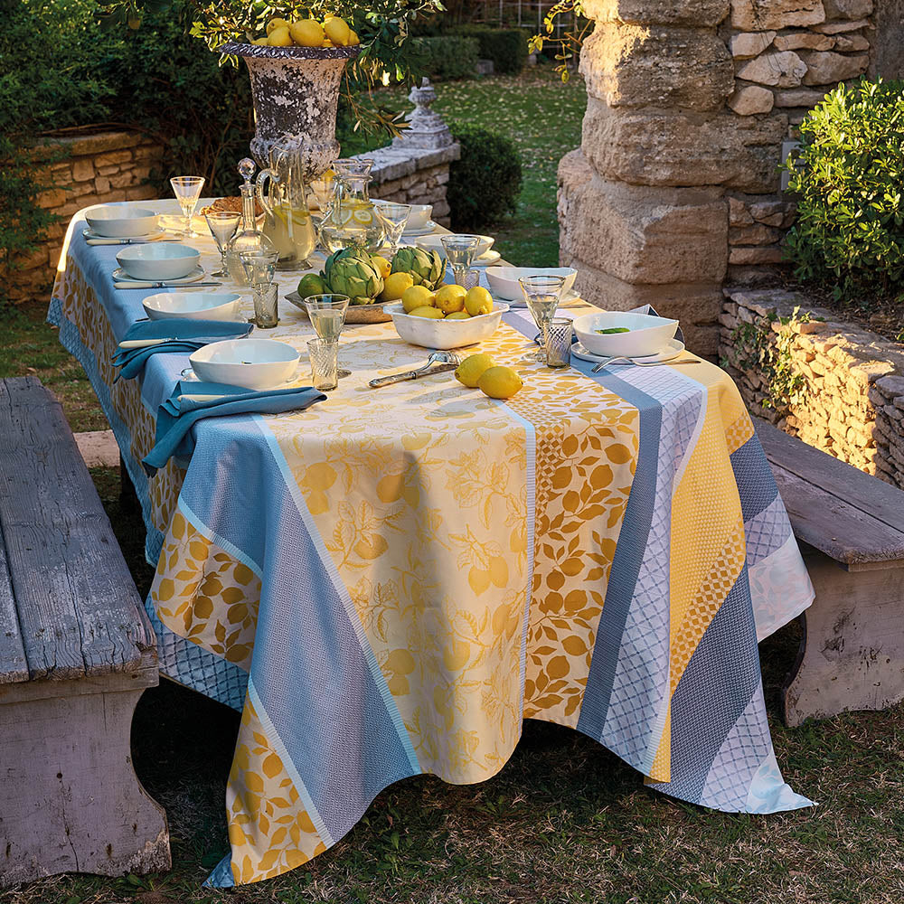 Mille Citrons Provence Jacquard Tablecloth in stripes of blue and yellow cotton with lemon motifs, styled in a rustic, outdoor dining environment with wood benches and matching table settings.