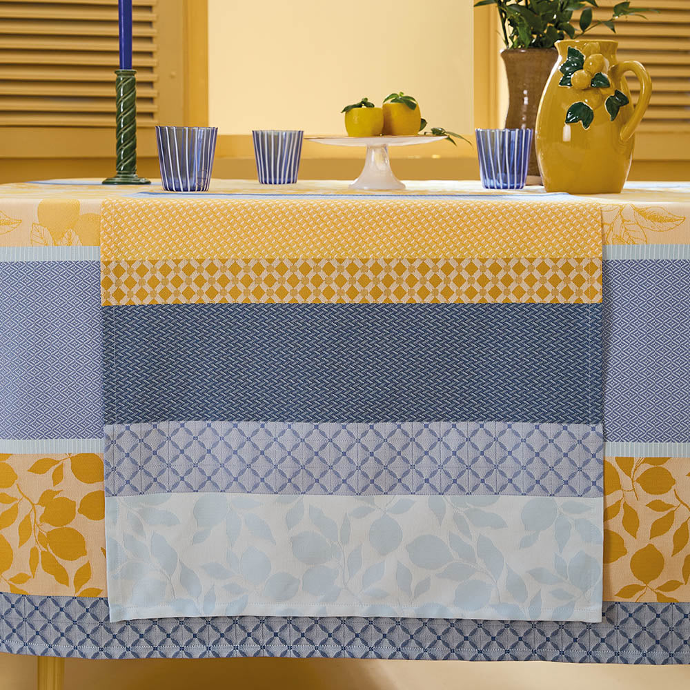 Mille Citrons Provence Jacquard Table Runner in striped blue and yellow cotton with geometric patterns draped over a dining table blue glasses and mustard yellow water pitcher.