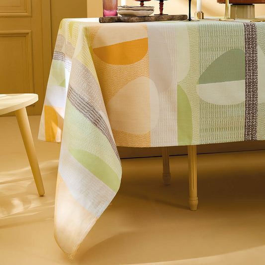Mille Galets Terre Cuite Tablecloth, cotton jacquard with yellow, brown, and green circular pebble motif, set for a relaxed dining scene in a soft yellow room.