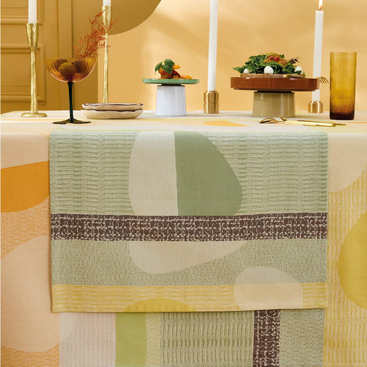 Mille Galets Terre Cuite Table Runner, yellow, brown, and green circular pebble motif on cotton jacquard, set on a yellow tablecloth.