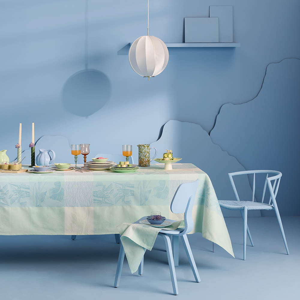 Mille Iris D'Eau Aqua Tablecloth, blue and white iris floral madras plaid on cotton jacquard, set for a garden-inspired dining scene in a sky blue room and matching chairs.