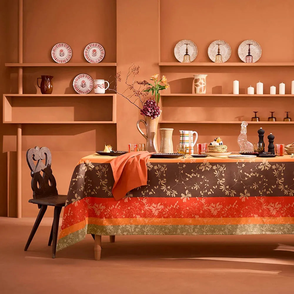 A woven cotton jacquard tablecloth featuring a warm, autumnal striped design with trailing leaves and floral motifs in yellow, brown, and orange, beautifully styled on a long dining table against a cozy terracotta wall with rustic wooden shelves.